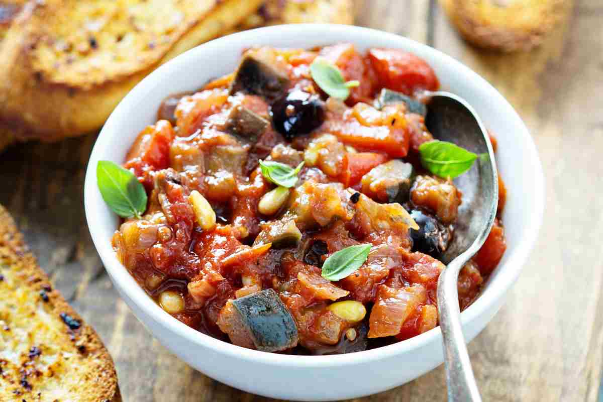 Caponata in una ciotola bianca e fette di pane abbrustolito sul tavolo